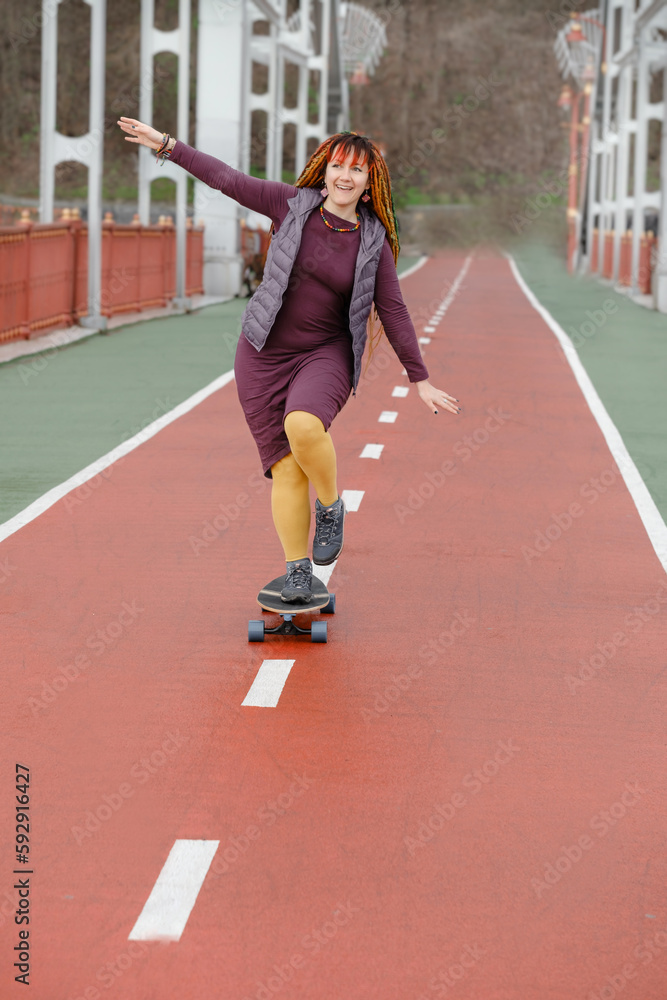 Obraz premium Funny free hipster Woman with dreadlocks on longboard on the road riding in the city at her 30s. female with bright colorful hair enjoy outdoor active lifestyle, play extreme sport