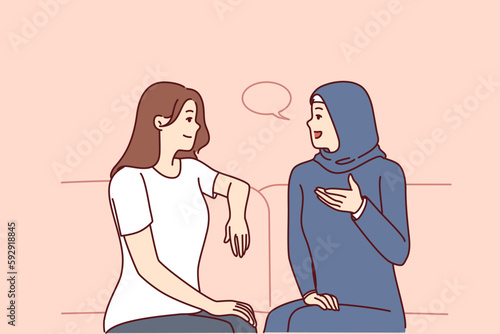Girls from different ethnicities are talking sitting on couch demonstrating tolerance and lack of cultural prejudice. Woman in muslim clothes is talking to girlfriend of european appearance 