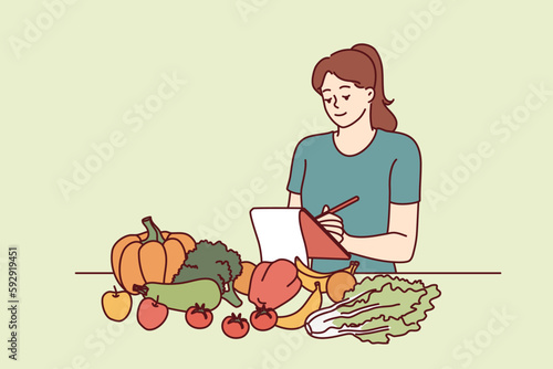 Woman nutritionist is standing near table with vegetables and making notes in notebook, making plan for new diet to detoxify body. Girl nutrition specialist uses checklist to mark diet ingredients 