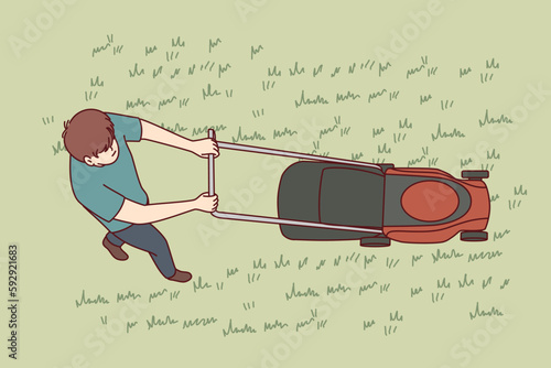 Man uses lawn mower to cut grass and wild weeds on golf course in park or in own backyard. Top view of young guy dressed in casual clothes with lawn mower working as gardener or janitor.