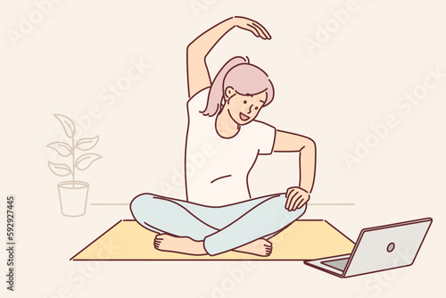 Woman sits on yoga mat and looks at laptop screen doing exercises during online broadcast