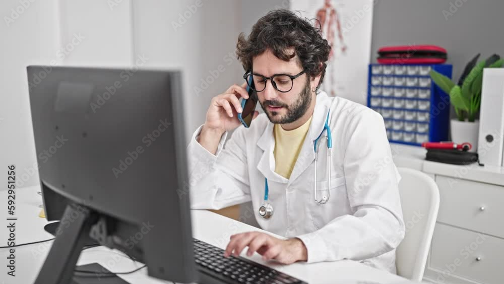 Young hispanic man doctor using computer talking on smartphone at clinic Young hispanic man doctor using computer talking on smartphone at clinic