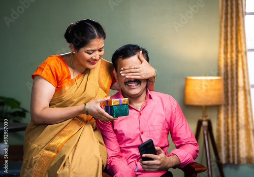 excited wife giving gift to husband by closing eyes while sitting on chair at home - concept of togetherness, happt long relationship and wedding anniversary. photo