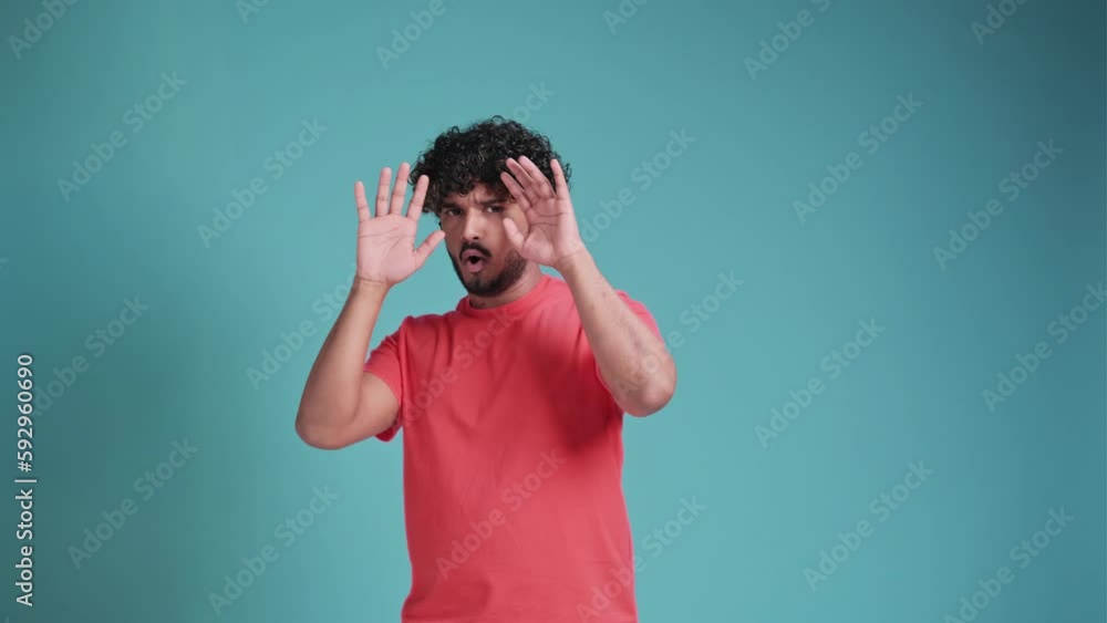 Horror, thriller, scare story. Portrait with young frightened indian man , guy screaming and raising hands with fear face in coral t-shirt on blue studio background. Human emotions, mood Horror, thriller, scare story. Portrait with young frightened indian man , guy screaming and raising hands with fear face in coral t-shirt on blue studio background. Human emotions, mood