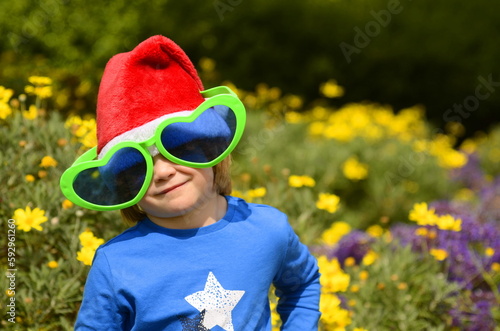 A little boy in a blooming garden in a Santa Claus hat. Concept: Christmas in summer, tropical holidays, traveling with children, snowless winter. © ShU studio