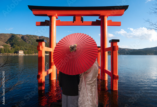Asian couple in kimono wedding dress standing togather with red umbrella and red torii gate background