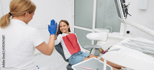 Lovely kid in dental chair with dentist satisfied after repairing teeth. Small girl giving high-five to dentist