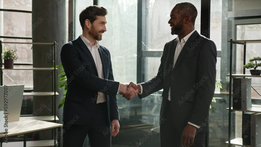 Two diverse multiracial businessmen business colleagues partners men handshaking in office posing smiling to camera Caucasian man and African American employee shaking hands agreement deal partnership Two diverse multiracial businessmen business colleagues partners men handshaking in office posing smiling to camera Caucasian man and African American employee shaking hands agreement deal partnership