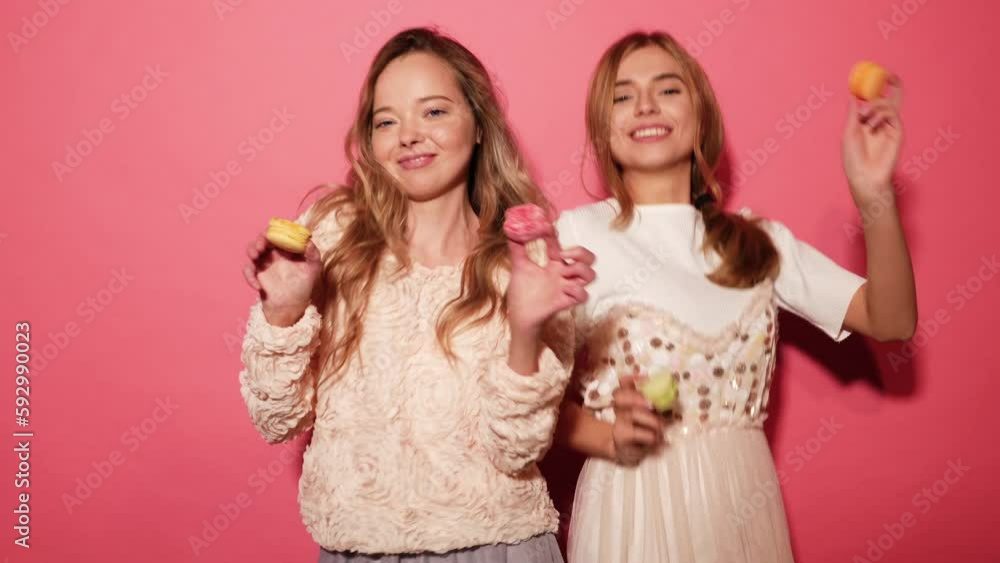 Two young charming beautiful smiling hipster girls in trendy summer clothes. Women eating macaroons, holding macarons. Posing on pink background Two young charming beautiful smiling hipster girls in trendy summer clothes. Women eating macaroons, holding macarons. Posing on pink background