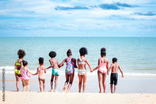 Group of African kids in swimsuit from behind have fun together on summer beach, boys and girls with black curly hair in line run to tropical sea water, happy childhood friends on holiday vacation.