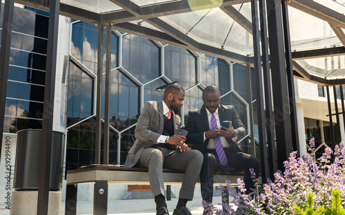 Two dark-skinned businessmen in suits communicate. Friends and business partners are sitting on a bench in the city against the backdrop of a glass building