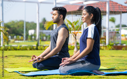 Side view shot of Indian young couple doing yoga or meditation at park during morning - concept of mindfulness, training and tranquility