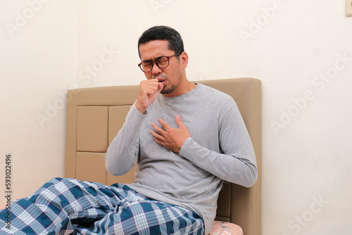 Adult Asian man sitting on his bed suffering bad cough