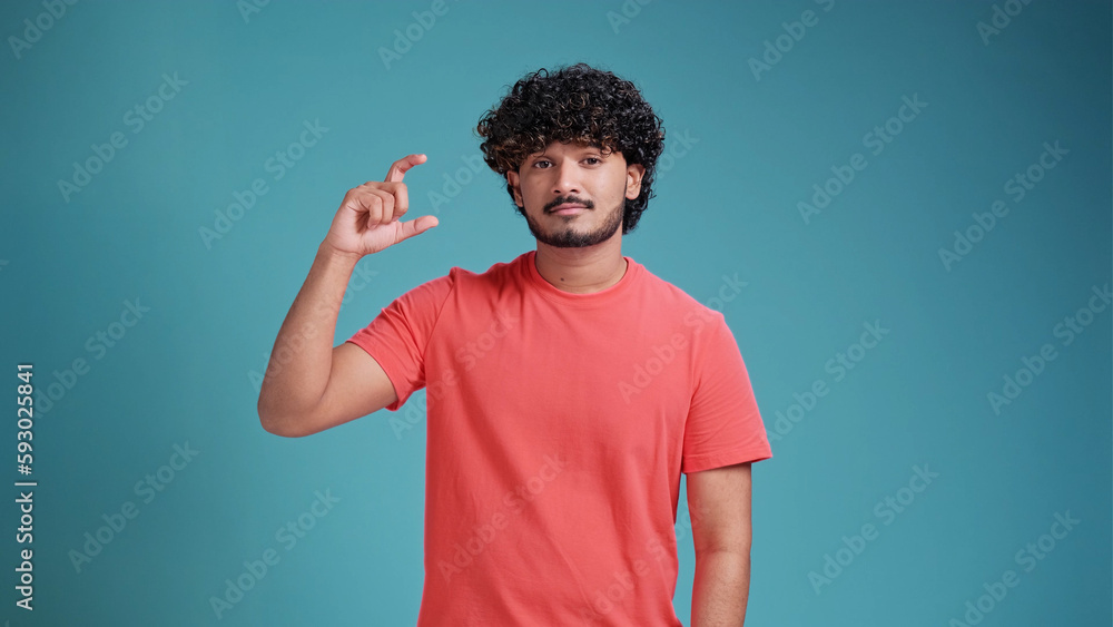 Disappointed millennial Indian man in glasses showing small size length with a pity in coral t-shirt on blue studio background, young guy looking at camera showing measurement with fingers. Disappointed millennial Indian man in glasses showing small size length with a pity in coral t-shirt on blue studio background, young guy looking at camera showing measurement with fingers.