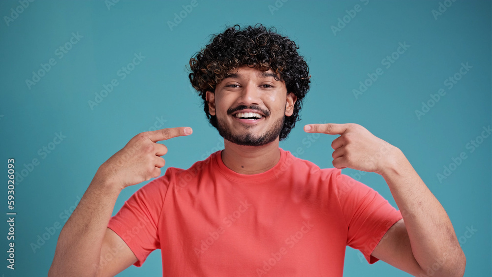 Happy confident Indian man with toothy smile pointing with fingers on his mouth looking at camera isolated on blue studio background. Health care, dental treatment concept. Happy confident Indian man with toothy smile pointing with fingers on his mouth looking at camera isolated on blue studio background. Health care, dental treatment concept.