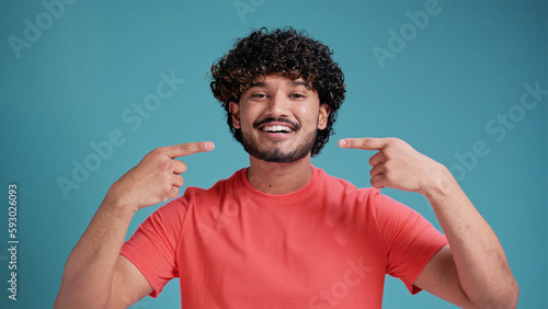 Happy confident Indian man with toothy smile pointing with fingers on his mouth looking at camera isolated on blue studio background. Health care, dental treatment concept.