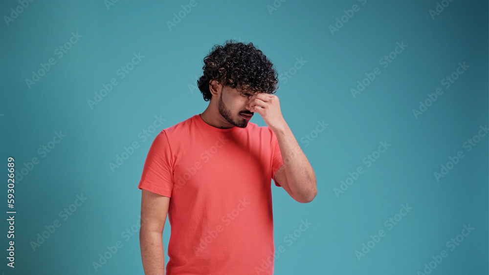 Young exhausted tired sad Indian manin coral t-shirt on blue studio background keep eyes closed rub put hand on nose isolated on plain pastel light blue cyan background studio portrait. Young exhausted tired sad Indian manin coral t-shirt on blue studio background keep eyes closed rub put hand on nose isolated on plain pastel light blue cyan background studio portrait.