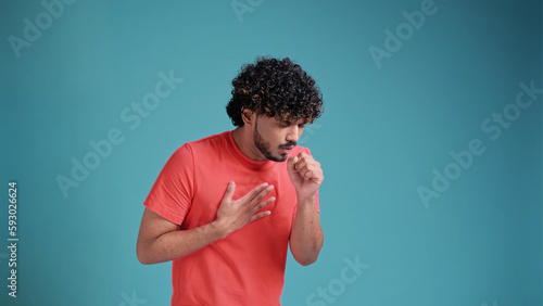 Young indian man in coral t-shirt on blue studio background, scarf sneezing or coughing covering mouth with hands