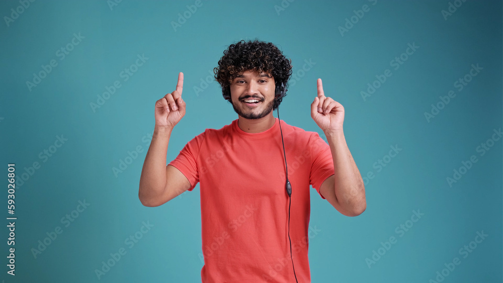 Young indian call center agent man using headset in coral t-shirt on blue studio backgroun pointing and showing with thumb up to the side with happy face smiling Young indian call center agent man using headset in coral t-shirt on blue studio backgroun pointing and showing with thumb up to the side with happy face smiling
