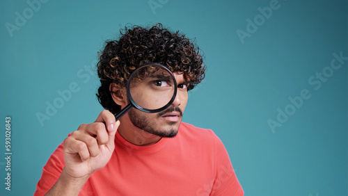 Funny Indian man looking through magnifying glass, searching or investigating something, standing in coral t-shirt on blue studio background