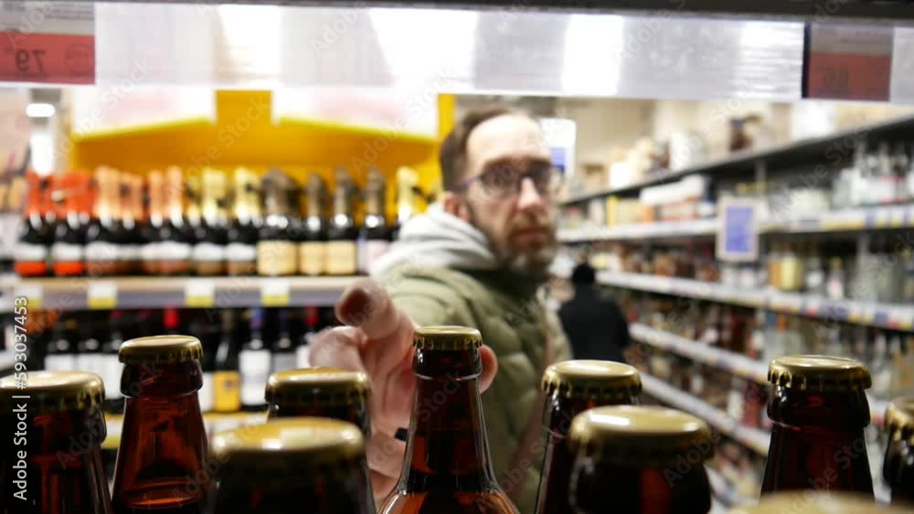 Many beer bottles on a store shelf close-up and a bearded man with glasses takes one