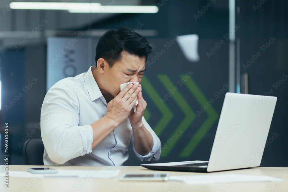 A young sick Asian man is sitting at work in the office. He wipes his nose from a runny nose with a napkin, coughs, feels bad. A young sick Asian man is sitting at work in the office. He wipes his nose from a runny nose with a napkin, coughs, feels bad.