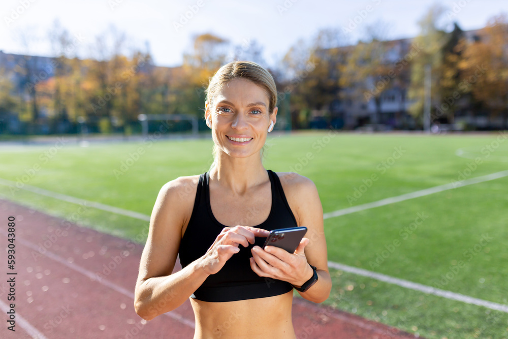 Young woman athlete runner standing in stadium with phone and headphones and tunes to listen to music, audiobook, podcast. Smiling at the camera.