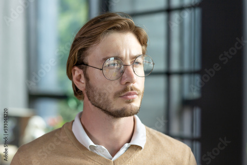 Close-up photo. Portrait of a young blond man in glasses looking seriously to the side, standing in an office, campus, wearing a shirt and a brown sweater.