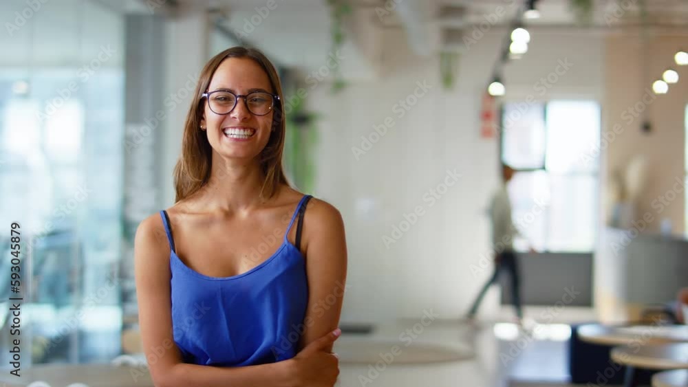 Camera tracks across smiling young businesswoman wearing glasses standing in modern open plan office looking into camera - shot in slow motion