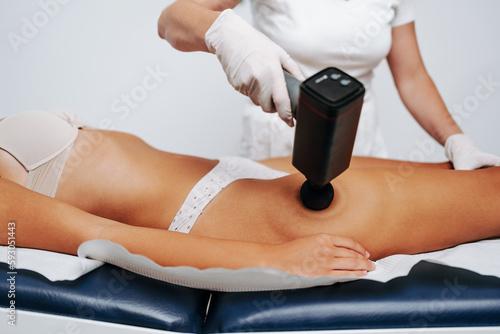 Young sporty caucasian woman having body massage using percussion pistol at beauty relax center. Electric massager pistol for special muscle treatment. Sports and pain recovery concept.