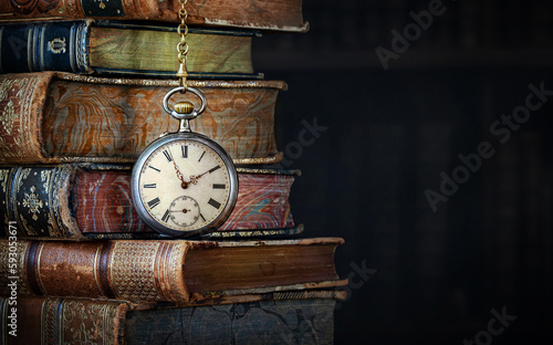 Old clock hanging on a chain on the background of old books.  Сlock as a symbol of time a books are a symbol of knowledge.. Concept on the theme of history, nostalgia, culture.