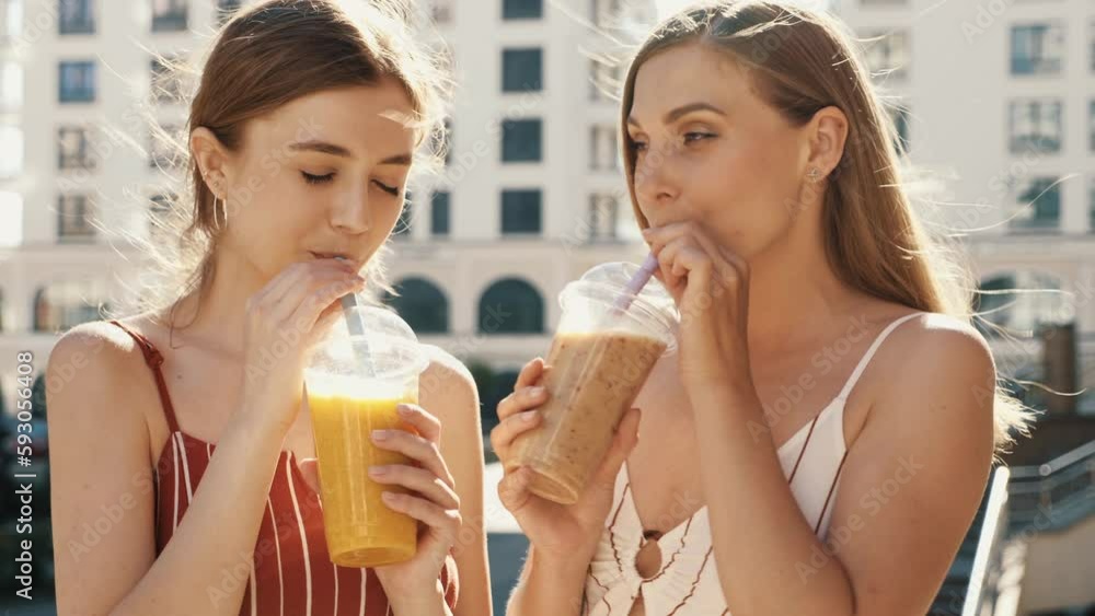 Two young beautiful smiling hipster female communicating and chatting. Carefree women posing outdoors. Positive models tasting each other fresh cocktail smoothie drink in plastic cup with straw Two young beautiful smiling hipster female communicating and chatting. Carefree women posing outdoors. Positive models tasting each other fresh cocktail smoothie drink in plastic cup with straw
