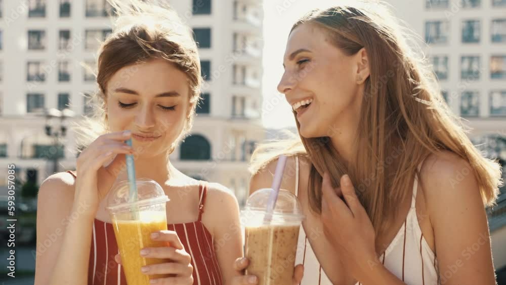 Two young beautiful smiling hipster female communicating and chatting. Carefree women posing outdoors. Positive models tasting each other fresh cocktail smoothie drink in plastic cup with straw Two young beautiful smiling hipster female communicating and chatting. Carefree women posing outdoors. Positive models tasting each other fresh cocktail smoothie drink in plastic cup with straw