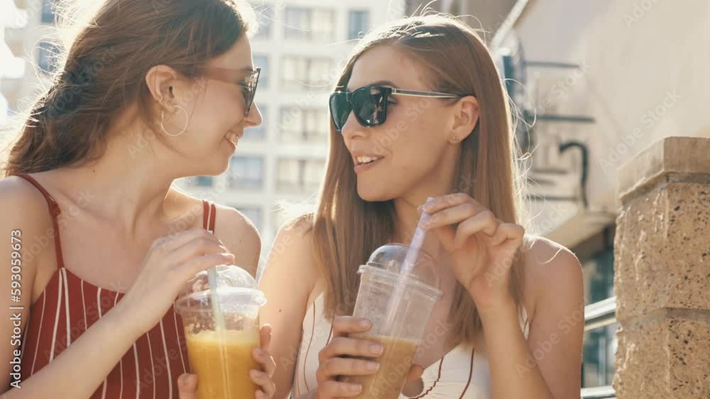 Two young beautiful smiling hipster female communicating and chatting. Carefree women posing outdoors. Positive models tasting each other fresh cocktail smoothie drink in plastic cup with straw Two young beautiful smiling hipster female communicating and chatting. Carefree women posing outdoors. Positive models tasting each other fresh cocktail smoothie drink in plastic cup with straw