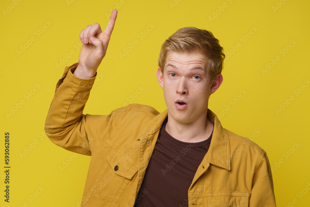 Amazed young man pointing up with his finger. Young 20s man wearing casual yellow clothes amazed and surprised looking and pointing with raised arm and finger above isolated on yellow background. Amazed young man pointing up with his finger. Young 20s man wearing casual yellow clothes amazed and surprised looking and pointing with raised arm and finger above isolated on yellow background.