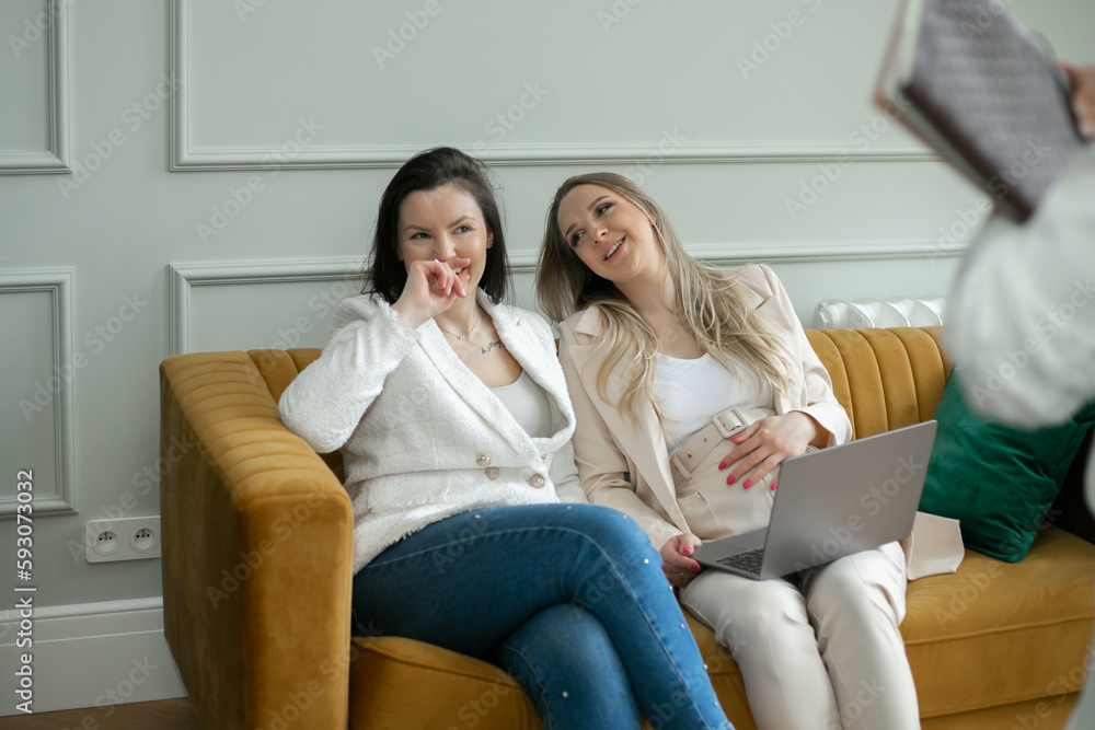 Happy women relax on sofa, work on laptop and joyfully talk in living room. Friends laugh and have fun together.