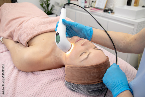 Relieved woman lying down in clinic salon while receiving laser facial beauty treatment, light radiofrequency technology