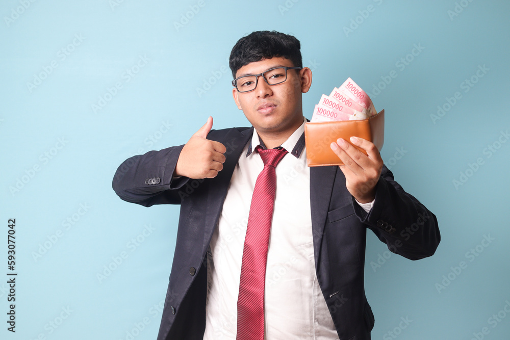 Portrait of young Asian business man in casual making thumb up while holding leather wallet with thousand rupiahs. Isolated image on blue background Portrait of young Asian business man in casual making thumb up while holding leather wallet with thousand rupiahs. Isolated image on blue background