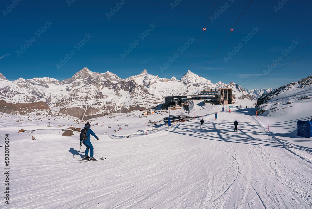 Rear view of skier skiing on snow covered landscape and scenic view of mountains and clear blue sky in background during sunny day at Zermatt, Switzerland, winter holiday travel concept
