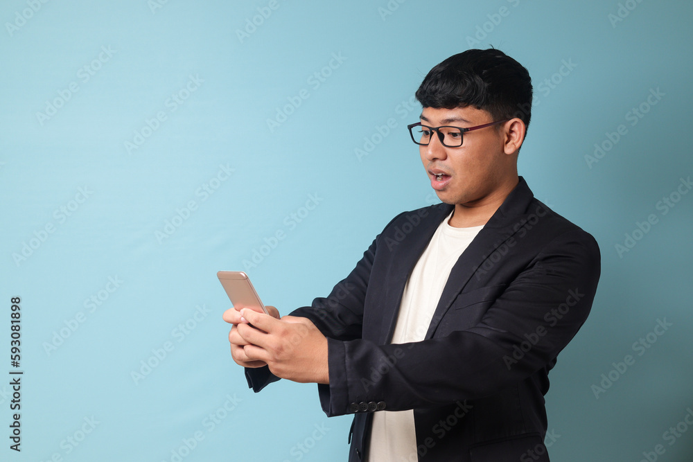 Portrait of young Asian business man in casual suit with surprised expression while looking at phone with both hands. Isolated image on blue background Portrait of young Asian business man in casual suit with surprised expression while looking at phone with both hands. Isolated image on blue background