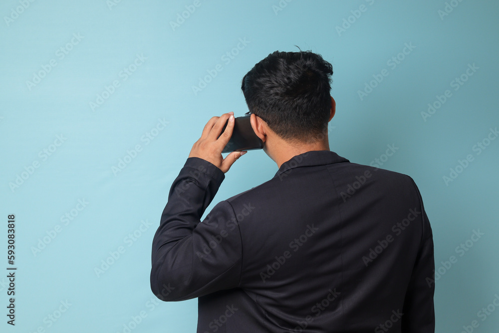 Portrait of young Asian business man in casual suit facing back while talking to phone. Isolated image on blue background Portrait of young Asian business man in casual suit facing back while talking to phone. Isolated image on blue background