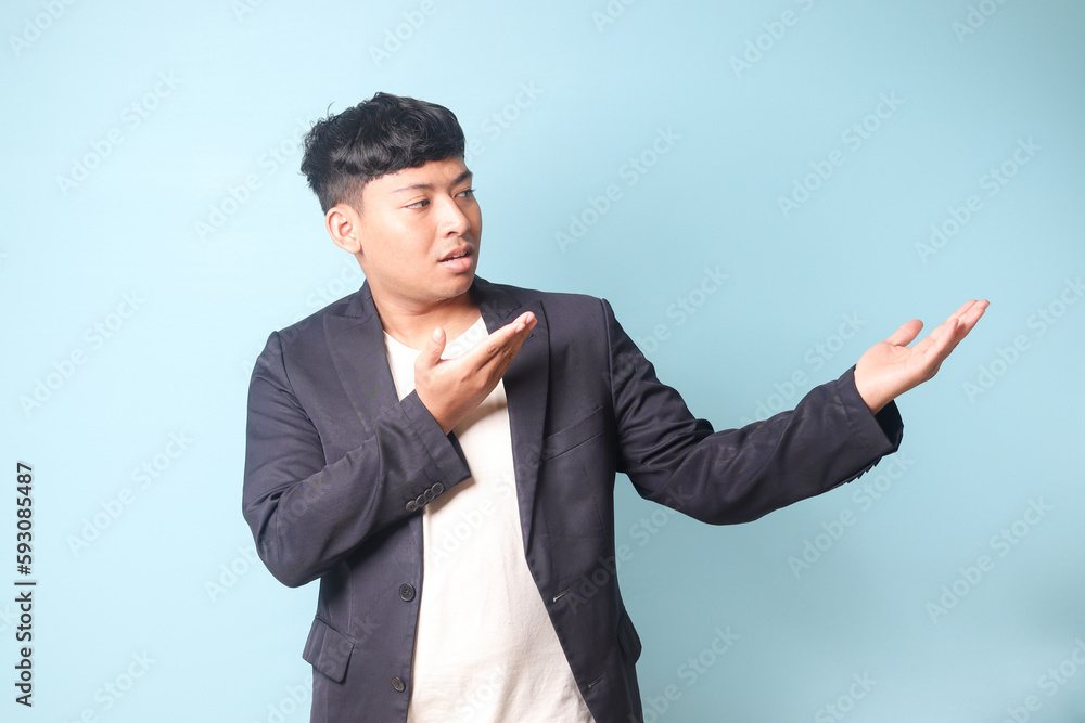 Portrait of young Asian business man in casual suit presenting something with both hands and looking at it. Isolated image on blue background Portrait of young Asian business man in casual suit presenting something with both hands and looking at it. Isolated image on blue background