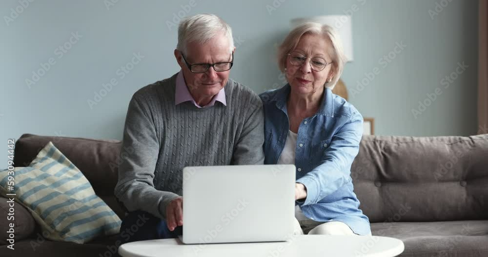 Elderly married couple talking at laptop at home, discussing booking service, buying goods on Internet stores, pointing at display, speaking, smiling, laughing, enjoying online communication Elderly married couple talking at laptop at home, discussing booking service, buying goods on Internet stores, pointing at display, speaking, smiling, laughing, enjoying online communication