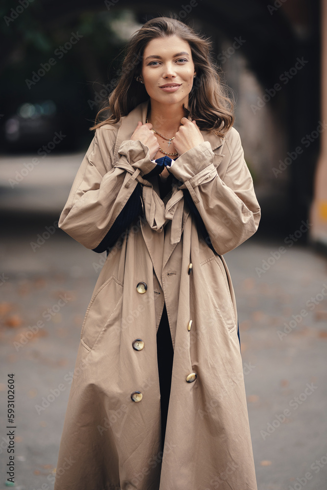 Fototapeta premium Elegant young girl in a beige raincoat posing outdoors