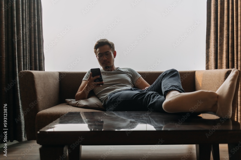 Asian man lies on sofa using phone with his feet on the table. Concept of manner