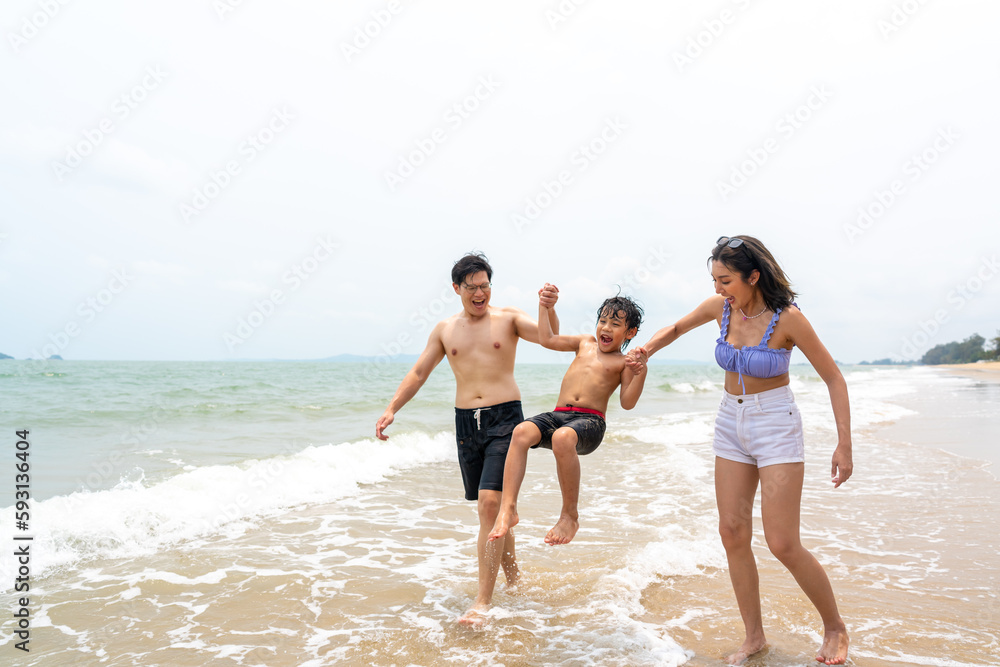 Happy Asian family travel ocean on summer holiday vacation. Parents and little son in swimwear have fun outdoor activity lifestyle walking and playing sea water together at tropical beach in sunny day