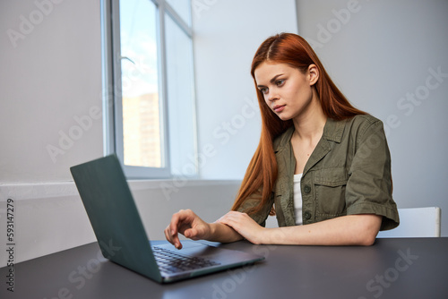 red-haired woman working in the office sitting by the window at the laptop