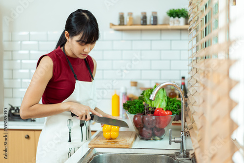 Happy cheerful beautiful Asian woman using cooking knife cutting a vegetables in kitchen. Woman preparing a vegan salad in kitchen.