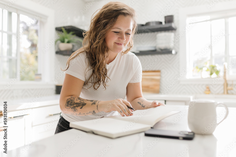 Happy Mid-Adult standing at kitchen counter journaling with coffee Happy Mid-Adult standing at kitchen counter journaling with coffee