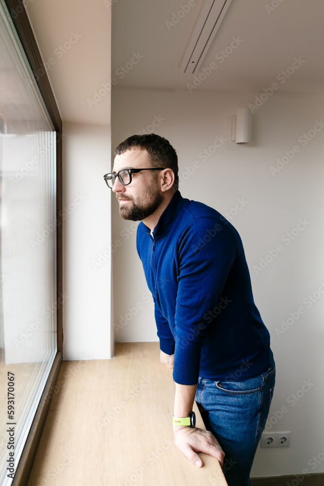 brunette man in glasses and a blue sweater looks out the window alone brunette man in glasses and a blue sweater looks out the window alone